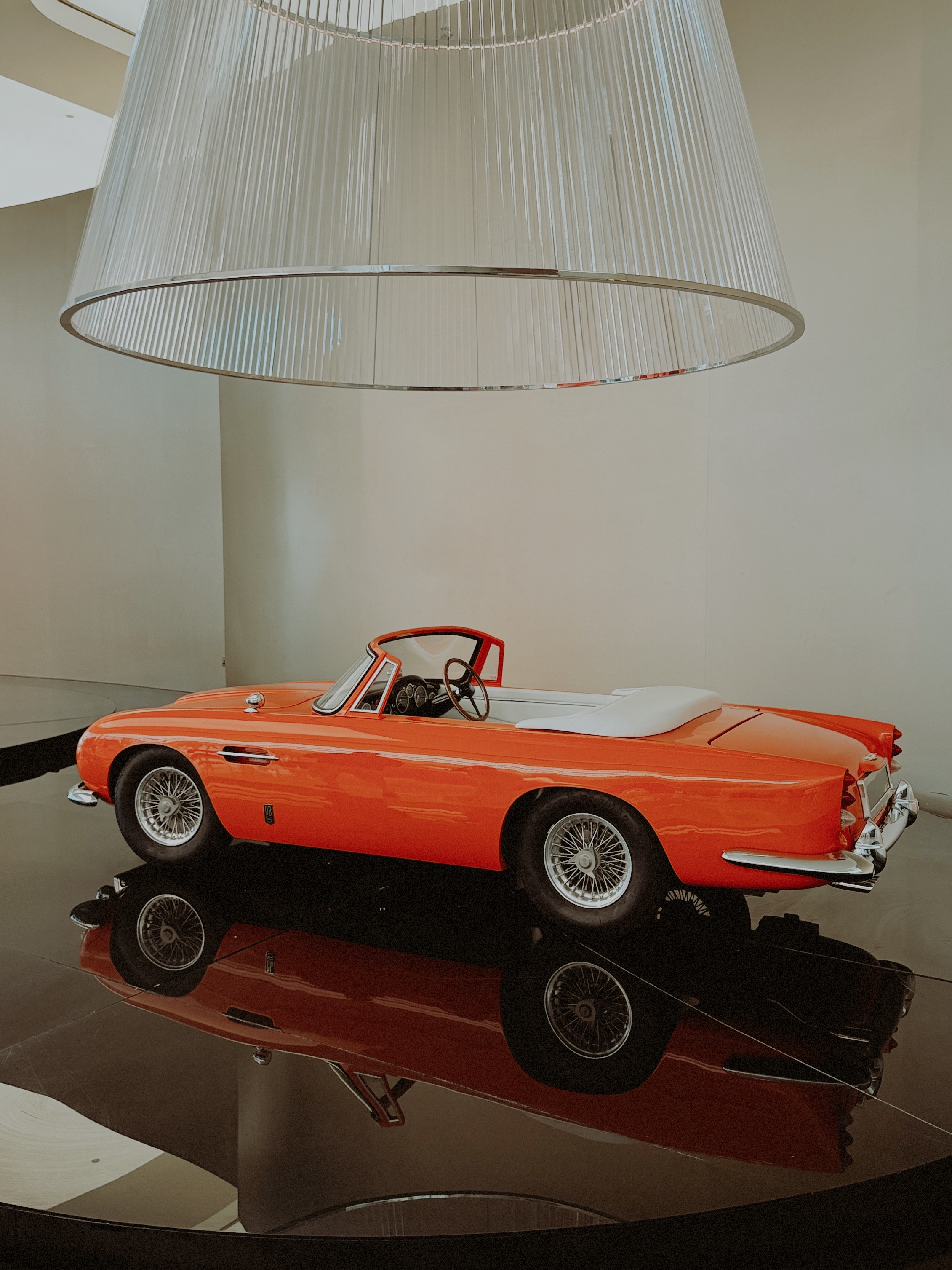 A vintage orange convertible beneath a vast glass-rod chandelier, reflected in a dark polished floor